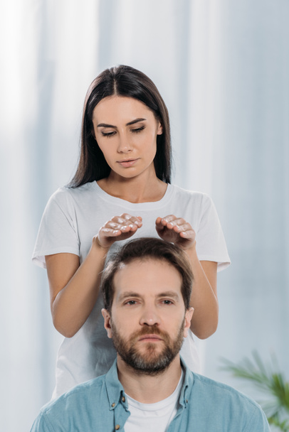 calm man looking at camera while receiving reiki treatment from young female healer  - Zdjęcie, obraz