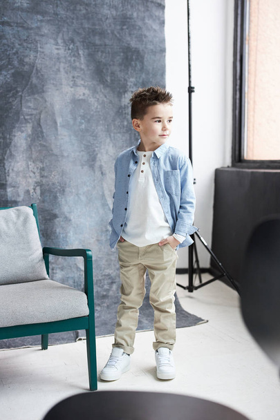 Stylish boy in blue shirt and beige pants walking in light loft studio with big window and grey chair - Photo, Image