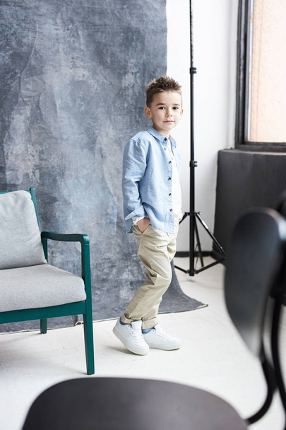 Stylish boy in blue shirt and beige pants walking in light loft studio with big window and grey chair - Foto, imagen