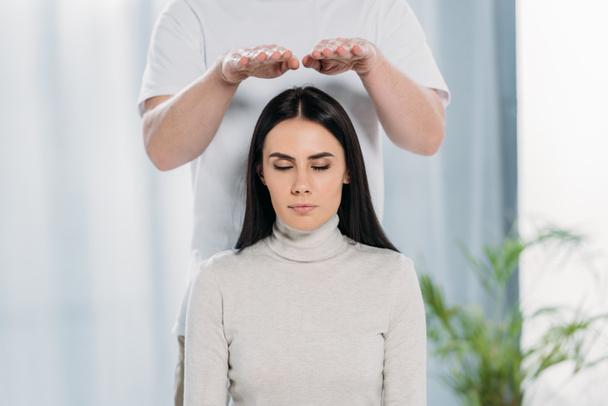 calm young woman with closed eyes receiving reiki healing treatment above head  - Foto, Bild