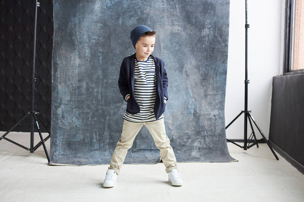 Stylish boy in blue shirt and beige pants walking in light loft studio with big window and grey chair - Foto, afbeelding