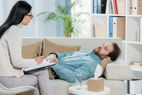 Junge Psychotherapeutin mit Brille schreibt auf Klemmbrett und sieht aufgebrachten bärtigen Mann auf Couch liegen - Foto, Bild