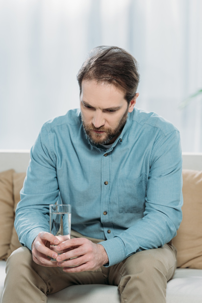 upset bearded man holding glass of water and sitting on couch in psychotherapist office - Photo, Image