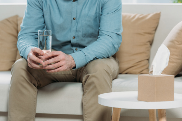 przycięte zdjęcie mężczyzny trzymającego szklankę wody, siedząc na kanapie w gabinecie psychoterapeuty - Zdjęcie, obraz