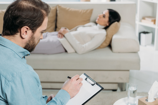 bearded psychotherapist writing on clipboard while upset patient lying on couch - Photo, Image
