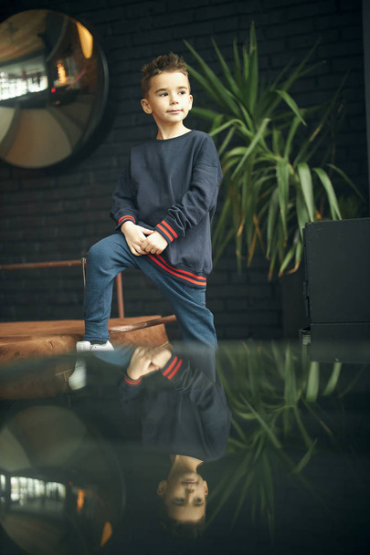 Fashionable boy sitting on chair near window in loft apartment, plant on backgroung. - Фото, изображение