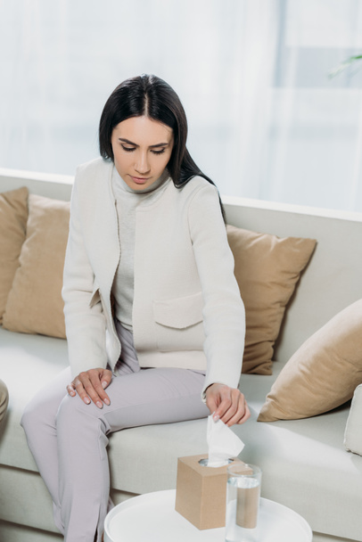 upset young woman holding paper tissue while sitting on couch in psychotherapist office - Photo, Image