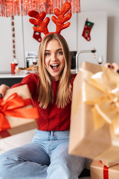 Retrato de una chica bonita de 20 años sentada en un sofá en la sala de estar con cajas de regalo mientras celebra la Navidad
 - Foto, imagen