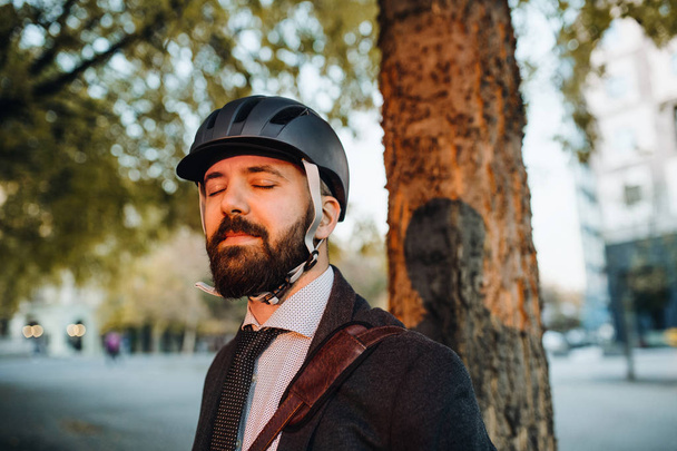 Businessman commuter with helmet standing in city at sunset, eyes closed. - Foto, imagen