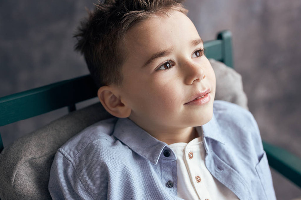 Close up portrait of little boy in blue shirt, sitting on chair, looking up - Fotoğraf, Görsel