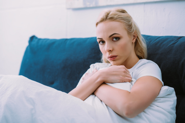 mujer deprimida sosteniendo una manta y mirando a la cámara mientras está acostada en la cama en casa
 - Foto, Imagen