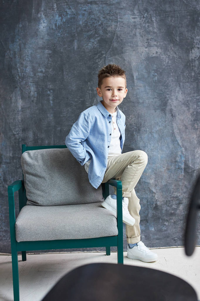 Fashionable boy in blue shirt  sitting on chair in loft studio, grey backgroung. - Фото, изображение