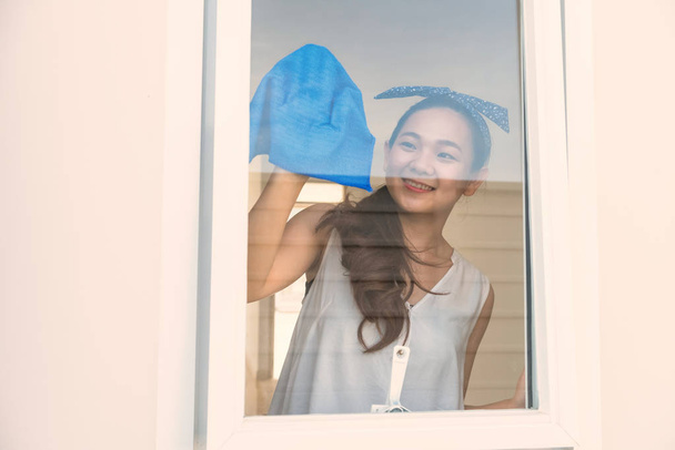 Mujer asiática feliz o ama de casa usando tela de microfibra azul para limpiar la ventana del nuevo hogar después de mudarse. Limpiar nueva casa para comenzar la vida familiar y comenzar el futuro. Primer día
. - Foto, Imagen