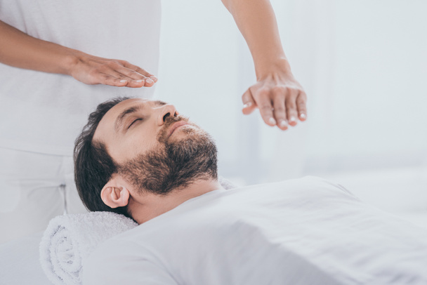 homem barbudo calmo deitado na mesa de massagem e mãos de curandeiro fazendo sessão de tratamento reiki
 - Foto, Imagem