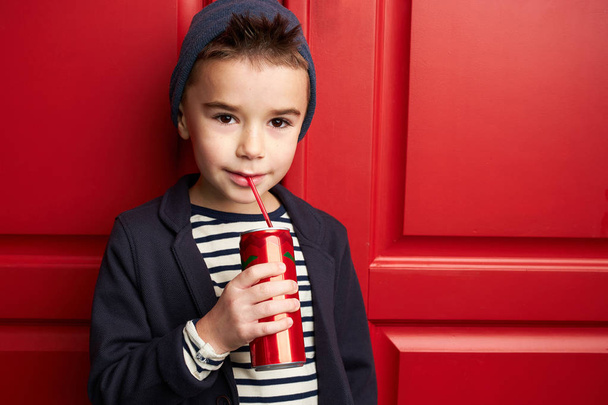 Little boy model posing in stylish wear drinking soda. Studio shot, red door on background. - Φωτογραφία, εικόνα