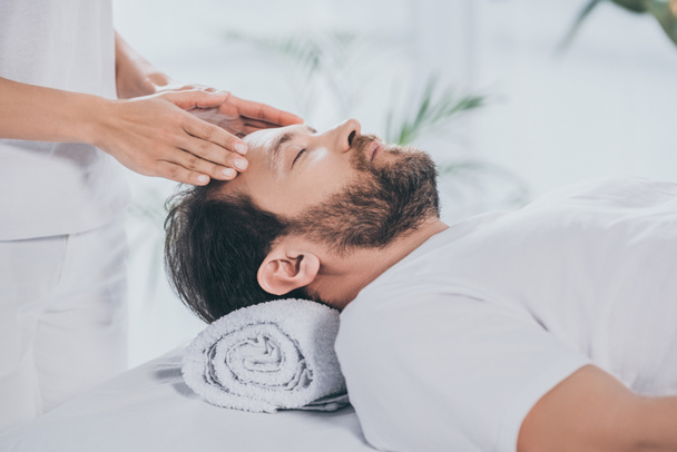 cropped shot of reiki specialist doing healing session to calm bearded man with closed eyes - Foto, Bild