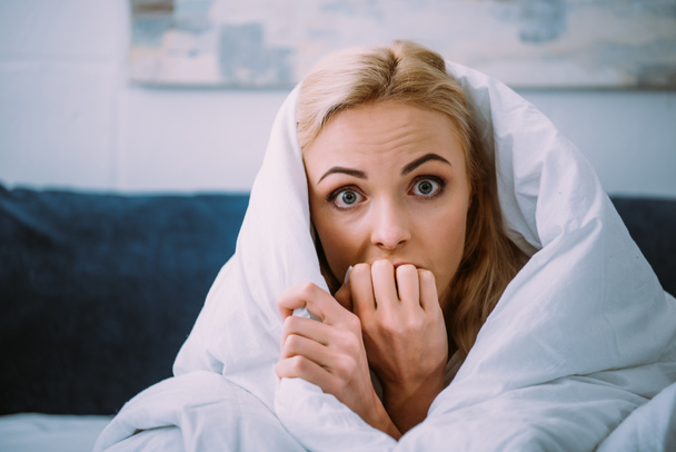 scared woman covered in blanket biting hand and looking at camera in bed  - Foto, imagen