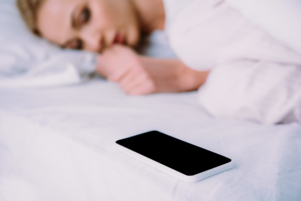 enfoque selectivo de teléfono inteligente con pantalla en blanco y mujer acostada en la cama sobre el fondo
 - Foto, Imagen