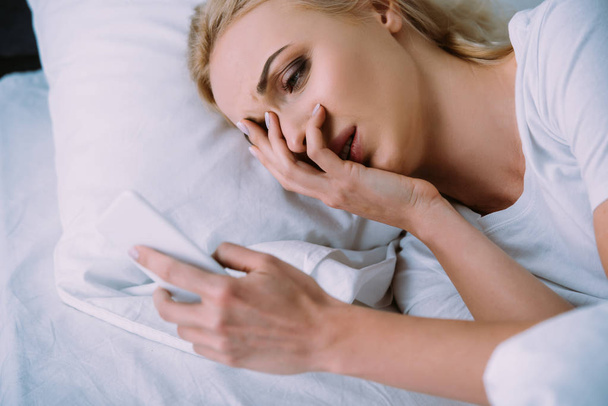 upset woman lying in bed, covering face with hand and using smartphone - Valokuva, kuva
