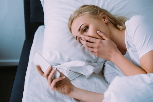 upset woman lying in bed, covering face with hand and using smartphone - Foto, afbeelding