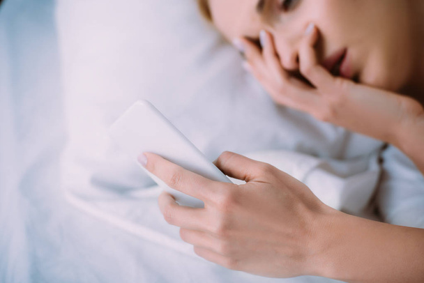 vue partielle de la femme couchée au lit, couvrant le visage avec la main et utilisant un smartphone
 - Photo, image