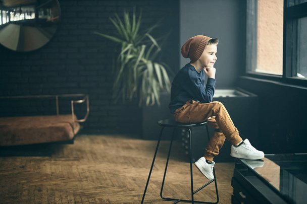 Fashionable boy sitting on chair near window in loft apartment, plant on backgroung. - Фото, зображення