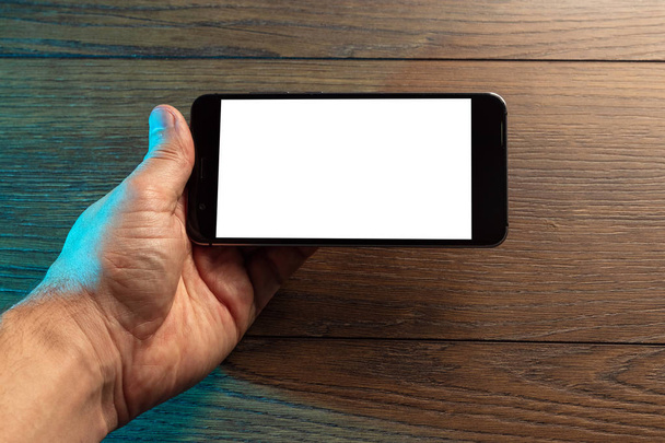 Closeup αρσενικό χέρια που κρατούν smartphone με λευκή οθόνη, το φόντο του ένα ξύλινο τραπέζι. Έννοια της κενό, κινητά παιχνίδια, κοροϊδεύω, διάταξη, αντίγραφο χώρου. - Φωτογραφία, εικόνα