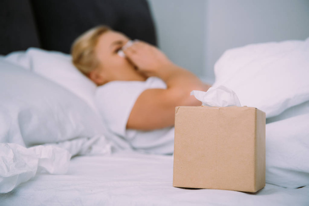 enfoque selectivo de la caja de tejido con la mujer limpiando lágrimas en la cama sobre el fondo
 - Foto, Imagen