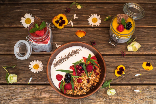 Breakfast with yogurt, granola and berries in bowl and jars of chia dessert on wooden table with flowers - Фото, изображение
