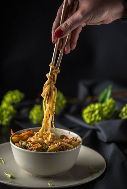 Female hand holding chopstick over bowl of asian seafood noodles on dark background - Zdjęcie, obraz
