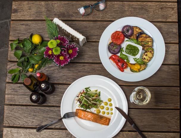 Dinner with piece of salmon, salad and grilled vegetables on plates on wooden table with glass of white wine - Foto, imagen