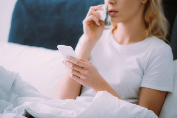 περικοπεί άποψη της γυναίκας σκουπίζοντας δάκρυα και χρησιμοποιώντας το smartphone στο κρεβάτι στο σπίτι - Φωτογραφία, εικόνα