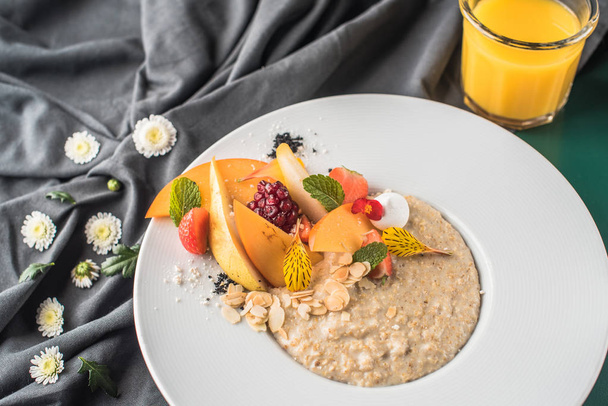Breakfast oatmeal with sliced fresh fruits on white plate with glass of fresh juice on black textile - 写真・画像
