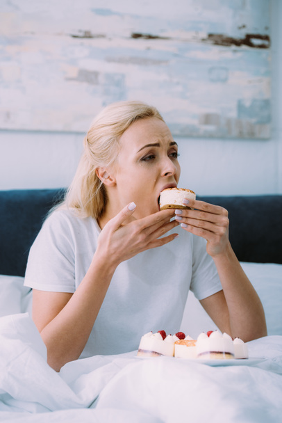 sad woman in pajamas eating sweet cake in bed alone  - Фото, изображение