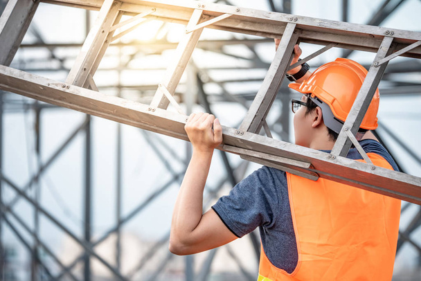 junger asiatischer Wartungsarbeiter mit orangefarbenem Schutzhelm und Warnweste, der auf der Baustelle eine Leiter aus Aluminium trägt. Bauingenieurwesen, Architekturbau und Haustechnik-Konzepte - Foto, Bild