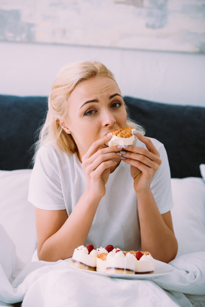 upset woman in pajamas looking at camera and eating cake in bed alone - Фото, зображення