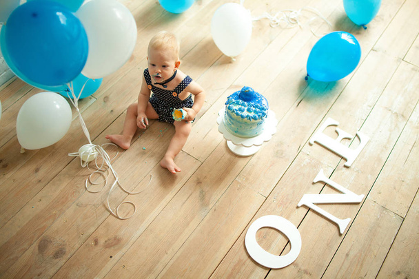 One year old boy is celebrating his first birthday, eating and smashing festive cake on background of blue balloons. Top view. - Photo, Image