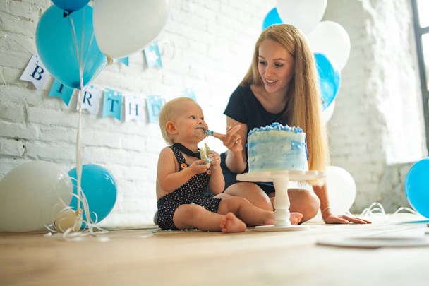 Mother is celebrating first birthday of her one year old son and feeding his by festive cake on background of blue balloons. - Foto, immagini
