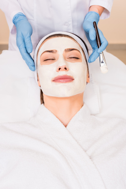kosmetička, stojící poblíž žena se zavřenýma očima a kosmetická maska na obličej v beauty salonu - Fotografie, Obrázek