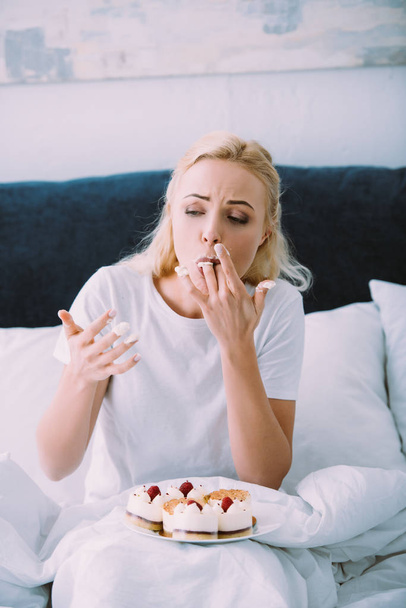 sad woman in pajamas licking fingers while eating cake in bed alone - Foto, Imagem