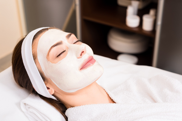 woman lying in white bathrobe with applied facial mask, smiling and closed eyes at beauty salon - Photo, Image