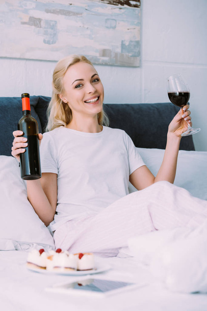 Kobieta szczęśliwa trzymając butelkę i kieliszek czerwonego wina podczas obchodzi urodziny w łóżku sam - Zdjęcie, obraz