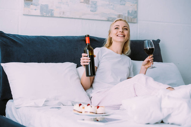 Lächelnde Frau mit Flasche und Glas Rotwein, während sie Geburtstag allein im Bett feiert - Foto, Bild
