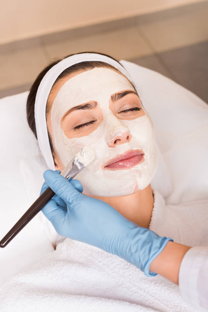 Kosmetikerin trägt im Schönheitssalon Kosmetikmaske auf Gesicht mit Kosmetikpinsel auf - Foto, Bild