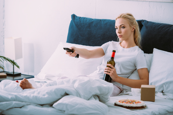 sad woman with wine bottle watching tv while celebrating birthday in bed alone - Photo, Image