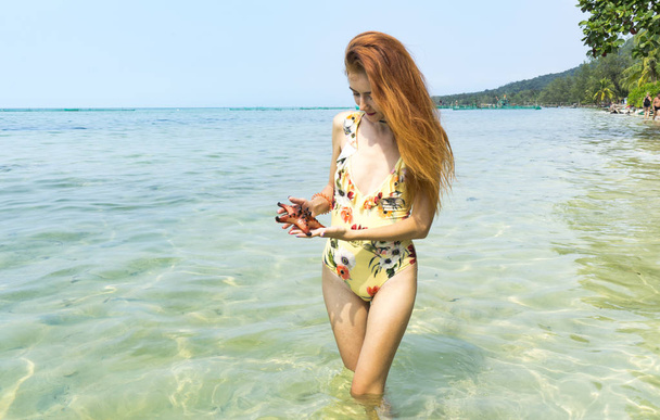 Red hair girl in bikini sitting with starfishes and holding coconut on the beach in Phu quoc island in a crystal clear sea, travel concept - Fotó, kép