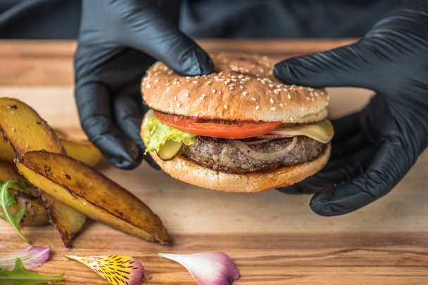 human hands in rubber gloves holding burger with fried potato wedges served on wooden board - Foto, immagini