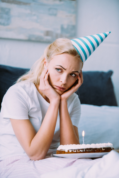 depressed woman with cake and party hat propping face with hands while celebrating birthday in bed alone - Photo, Image