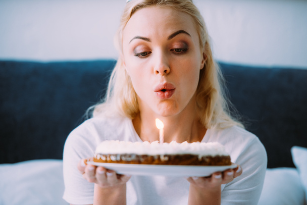selective focus of woman blowing out candle on cake while celebrating birthday in bed alone - Photo, Image