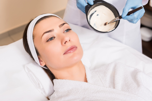 attractive woman lying in bathrobe and hairband while beautician mixing face mask at beauty salon - Foto, afbeelding